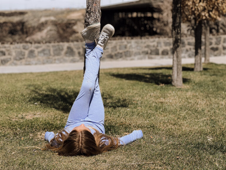 A young girl lays on the ground and props her legs up on a tree trunk.