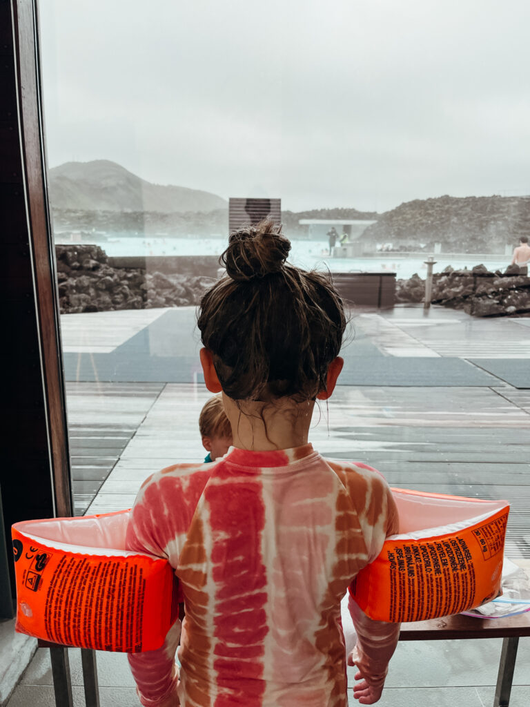 A young girl has her back to the camera and looks out the glass windows of the Blue Lagoon. There are black lava rocks in the background and silica water at this kid-friendly Icelandic geothermal pool.