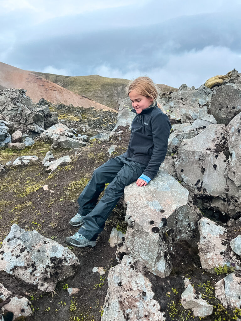 A young girl leans against lava rocks. She is wearing all black hiking clothes and has grey hiking boots on.