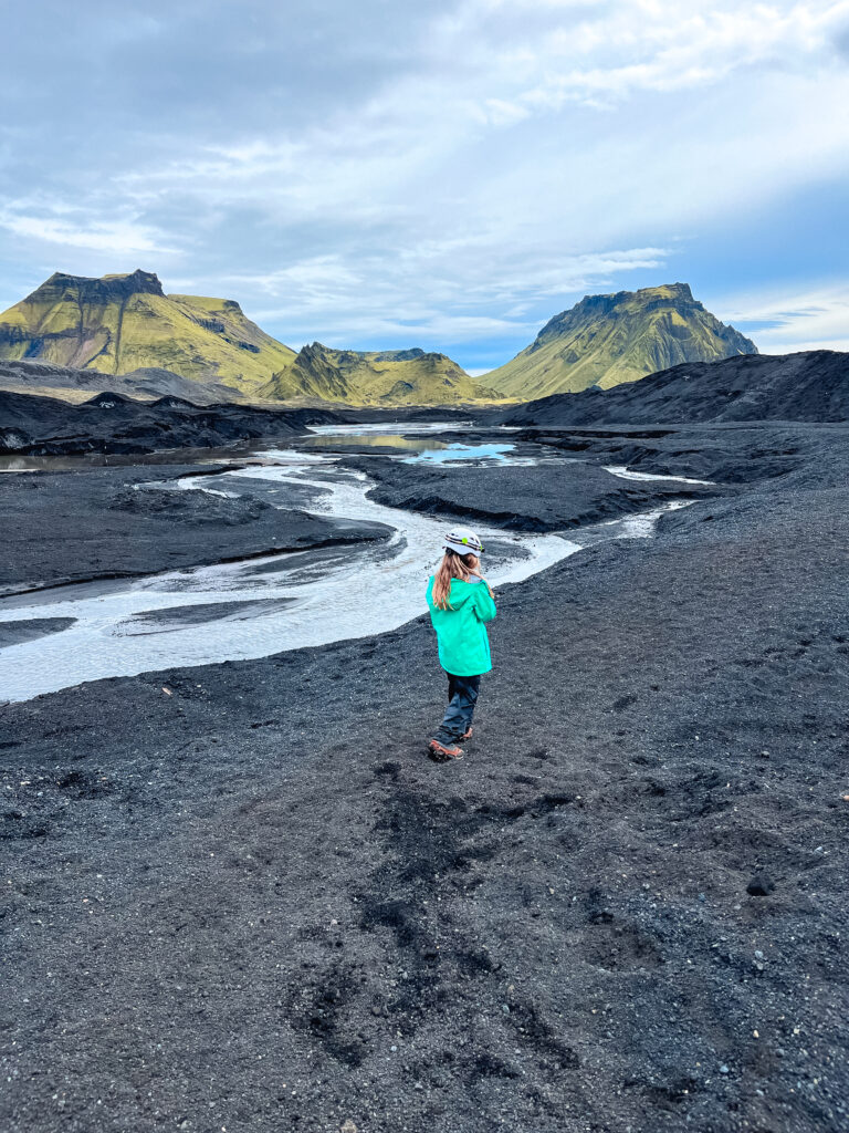 A young girl walking through silt ash on the katla glacier with a glacial river and mountains in the background.