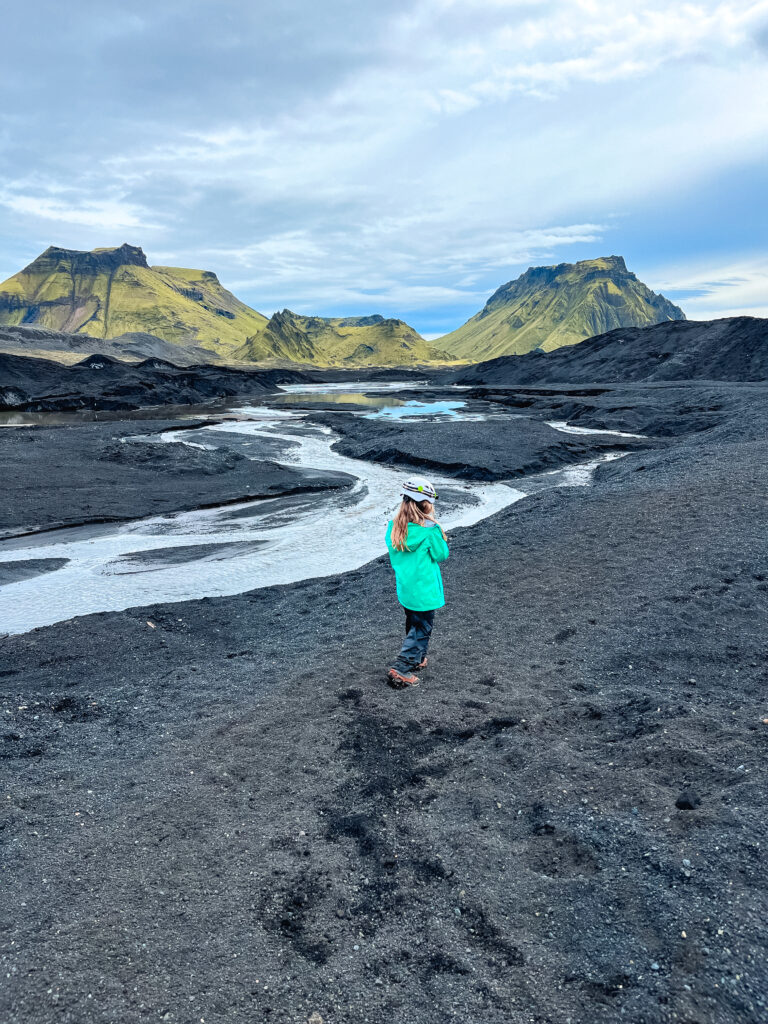 A young girl walks on volcanic sand in front of green mountains and glacial river on one of the most kid-friendly hikes in Iceland.