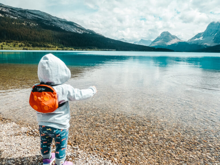 A young girl throws rocks in a glacial lake while wearing a toddler size backpack.