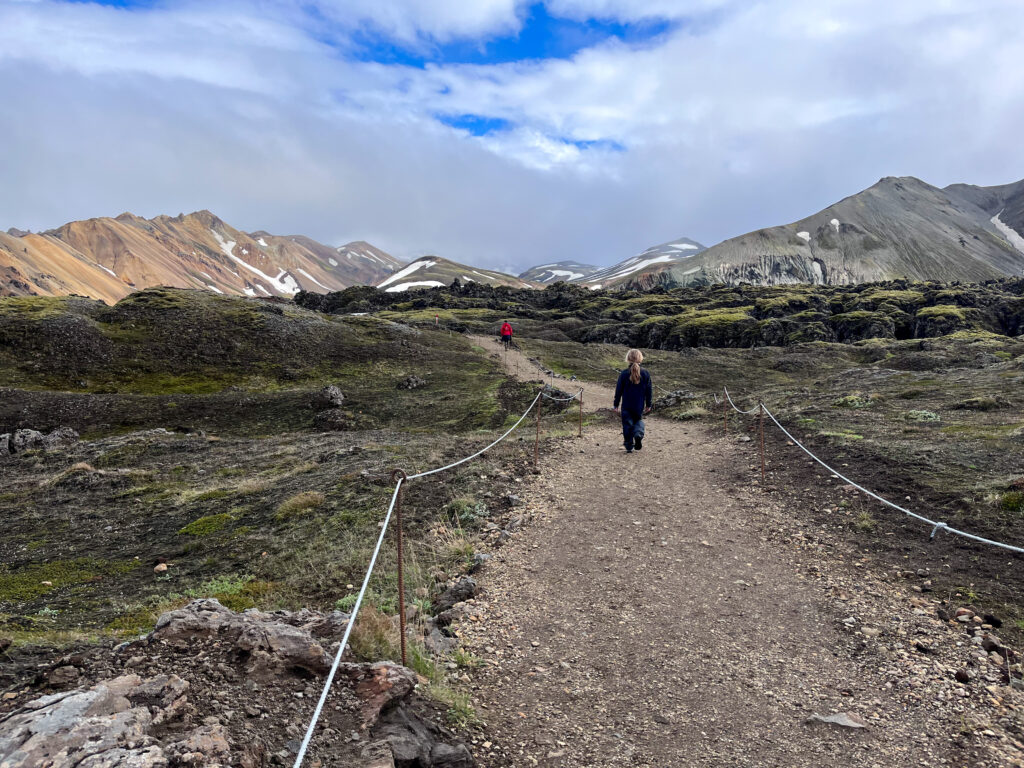 A young girl in all black walks along a dessert dirt trail. The painted mountains of Iceland are on either side of her and in the background.