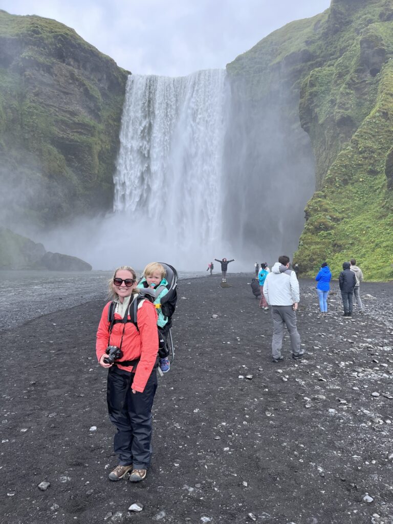 A woman in a bright orange rain jacket is wearing a toddler in a hiking carrier in front of Skogafoss waterfall.