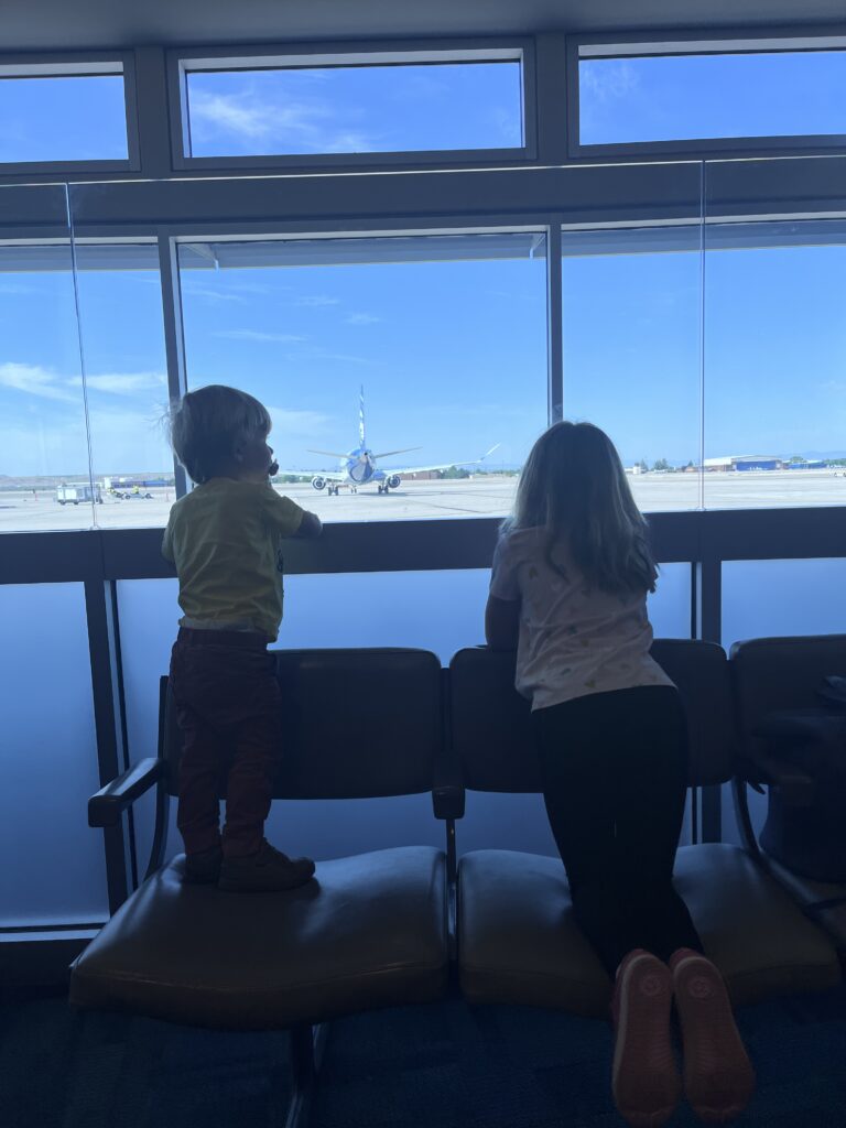 Two children stand on seats in an airport looking out the window at airplanes.