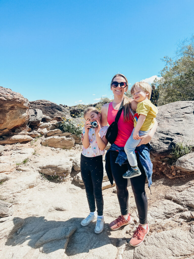 A woman holds a toddler and has her arm around a young girl smiling on a rocky trailhead, the Mormon trail, one of the best Scottsdale activities for kids.