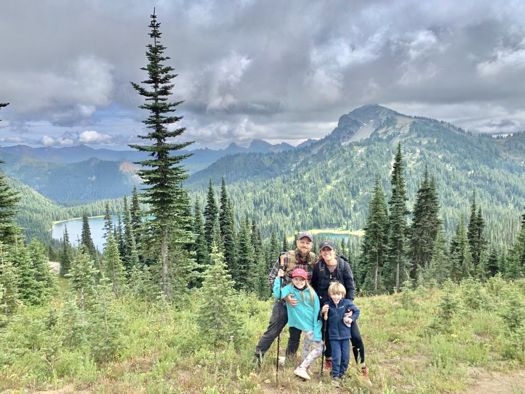 adventure mom priscila poses with her husband and young children in front a green mountain range.