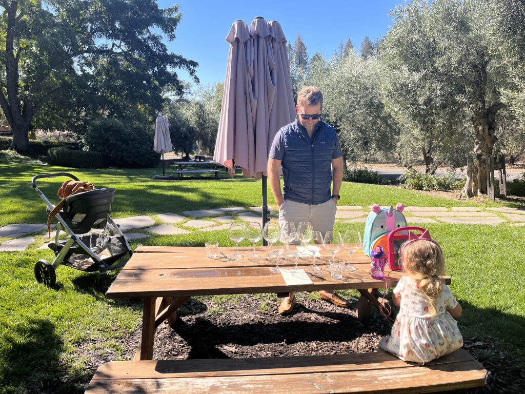 A man is looking at wine glasses for a wine tasting while a young girl watches and ipad and a baby naps in a stroller.