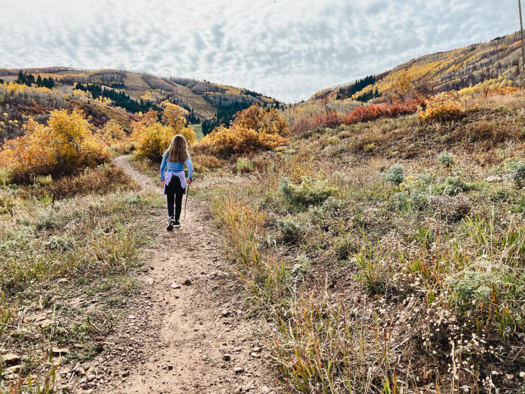 A young girl hikes on Jenni's Trail in Park City in October. Yellow and red trees dot the hillside in front of her.